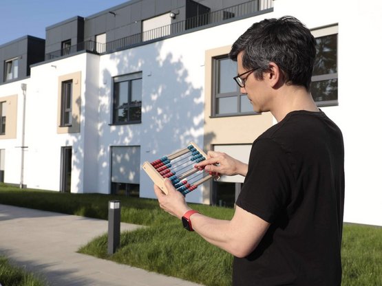 Ein junger Mann steht mit einem Rechenschieber vor einer Reihe Einfamilienhäuser und berechnet etwas.